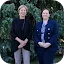 Two smiling women in professional attire stand side-by-side in front of a evergreen blue spruce tree. The woman on the left wears a black top and light-colored pants. The woman on the right wears a navy blue double-breasted blazer and silk top. They are featured as speakers for the Google and The Aspen Institute dialogue on 'How AI is changing the cybersecurity landscape'.
