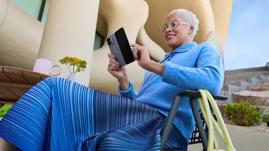 A person wearing an all blue outfit is sitting at a table smiling while using their Google Pixel 10 Pro Fold.
