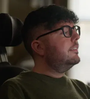 A side profile of a man wearing glasses and a green sweater, seated in a high-back chair near a window.