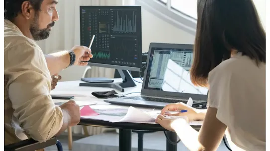 A man in a wheelchair and a woman are collaborating in a modern, sunlit office. They are discussing data visualizations displayed on a desktop monitor and a laptop. The man points toward the screen with a pen, emphasizing the human-centric approach to analyzing AI-generated insights in a workspace designed for accessibility.