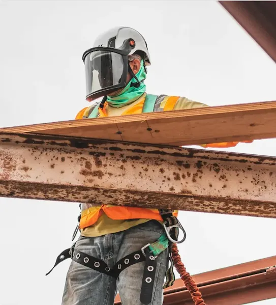 A construction worker wearing a safety helmet with a face shield, high-visibility vest, and fall protection harness, standing on a steel structure while holding a wooden plank.