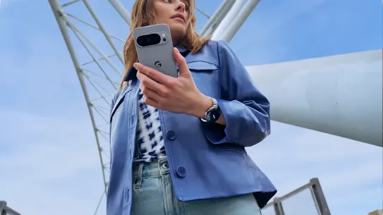 A person wearing a blue leather jacket and jeans is holding a Moonstone Google Pixel 10 phone underneath a structure outside.