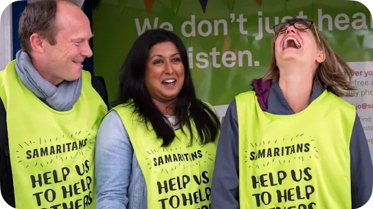 Three cheerful volunteers wearing high-visibility lime green vests that read "Samaritans Help Us To Help Others" stand side-by-side, sharing a laugh in front of a green banner.