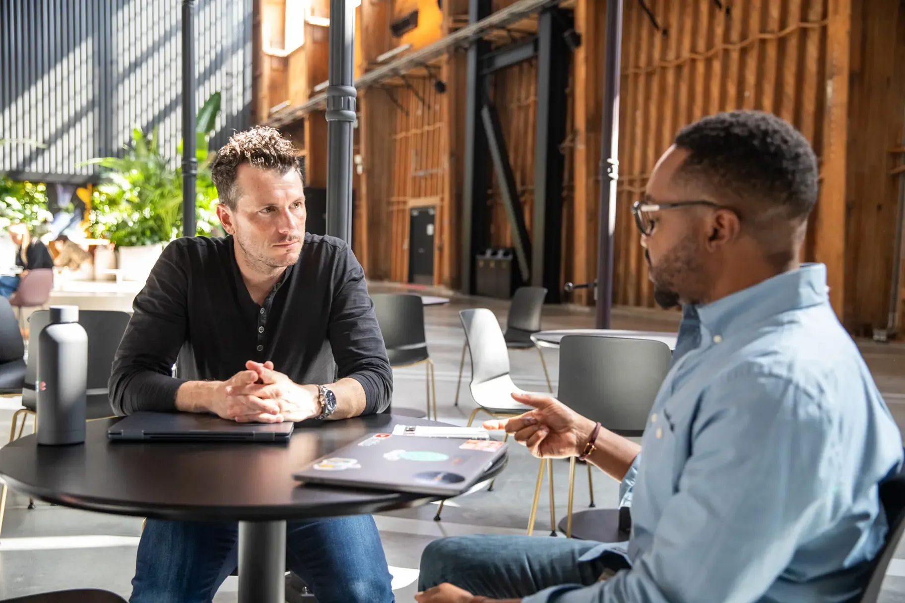 Two men sitting at a table talking to each other in the common space of an office. 
