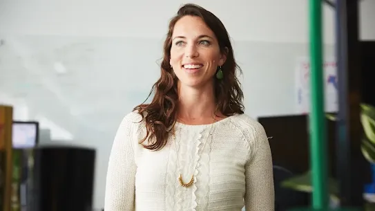 A woman with long, wavy brown hair and green eyes smiles warmly while looking off-camera. She is wearing a white knit sweater, green drop earrings, and a gold necklace, standing in what appears to be a bright office environment.