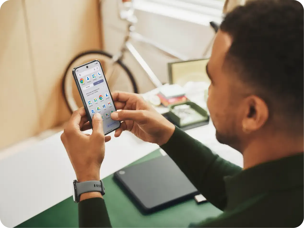 A man in a green shirt is holding his Pixel 9 in front of his face while seated at his desk.