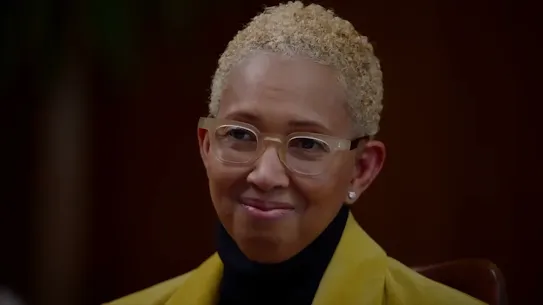 A headshot portrait of Ivor Horn, Chief Health Equity Officer and Director at Google, smiling slightly. She has short, blonde curly hair and is wearing translucent-framed glasses, a black turtleneck, and a yellow blazer against a soft, dark background.