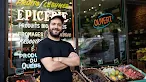 A smiling man with a beard and a black cap stands with his arms crossed in front of a grocery store window displaying French text and an "Ouvert" sign, with baskets of fresh fruit in the foreground.