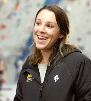 A smiling woman wearing a black hooded jacket with a Black Diamond and Momentum Climbing logo, looking to the side inside an indoor climbing gym with a blurry climbing wall in the background.