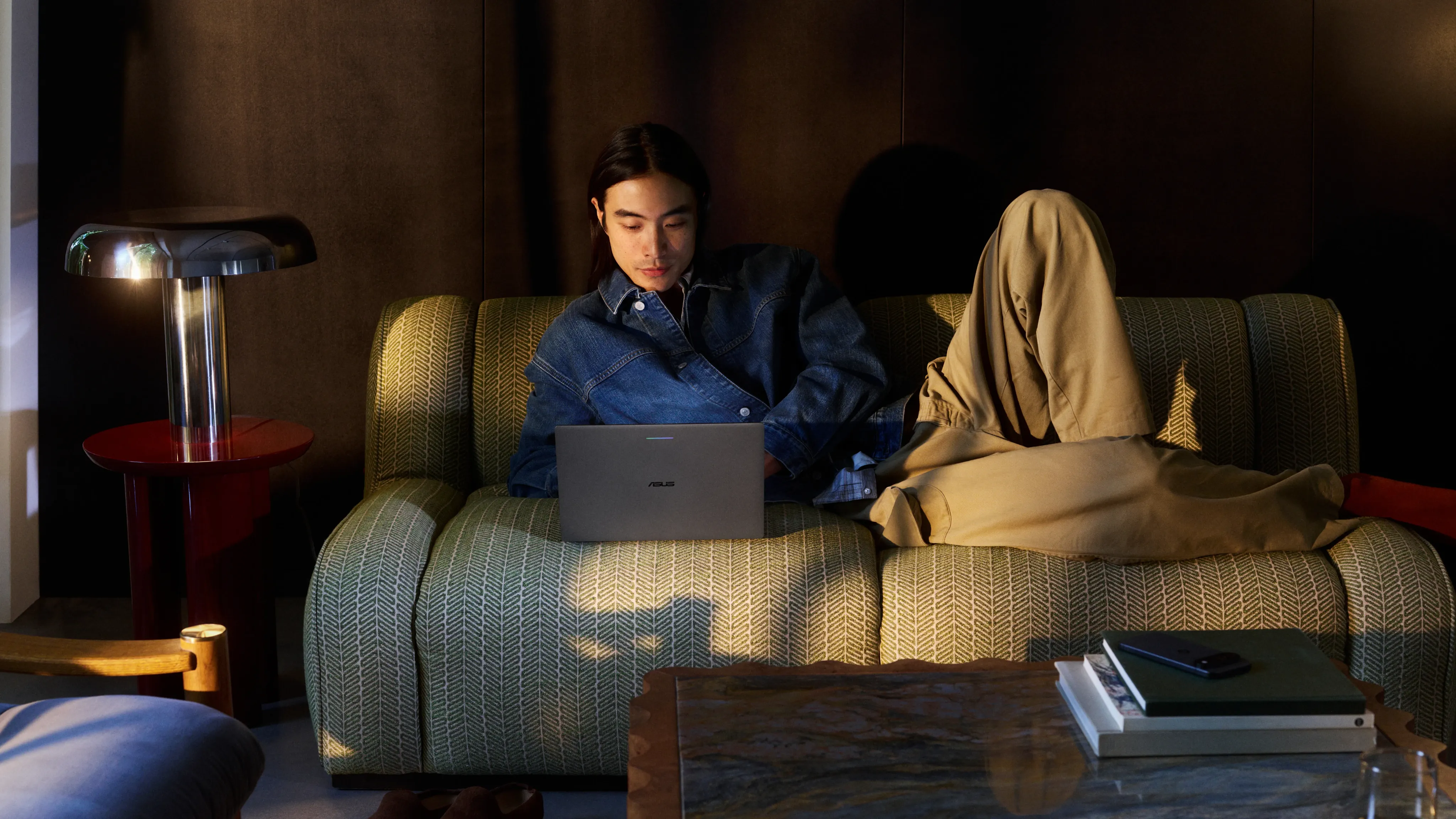 A person in a denim jacket relaxing on a patterned sofa, using a laptop, with a modern table lamp beside them.