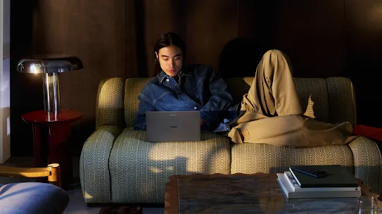 Una persona con una chaqueta de mezclilla se relaja en un sofá estampado, usa una laptop y tiene una lámpara de mesa moderna a su lado.