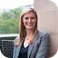 Professional headshot of a smiling woman with long strawberry-blonde hair, wearing a navy and white patterned blazer and a dark top. She is seated on a black wicker chair on an outdoor terrace. Featured speaker for the Google and Center for Democracy and Technology (CDT) dialogue: 'What’s Driving Optimism around AI in 2024'.