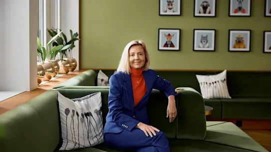 A professional portrait of a blonde woman in a blue suit and orange turtleneck, sitting on a green velvet sofa in a modern office. Behind her is a green wall decorated with framed whimsical animal portraits. The setting is bright and warm, appearing in the "Foundation of Innovation" report for the EU.