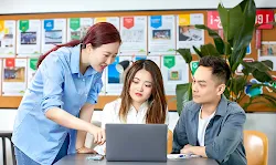 Two women and one man are discussing in room, looking on screen of laptop.