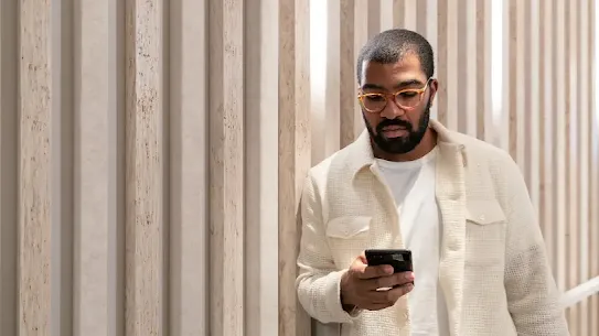 A man with a beard and glasses stands against a wall with vertical wooden slats. He is wearing a white t-shirt, a cream-colored textured button-down shirt, and black pants. He is looking down at a smartphone in his hands and holding a laptop under his arm.