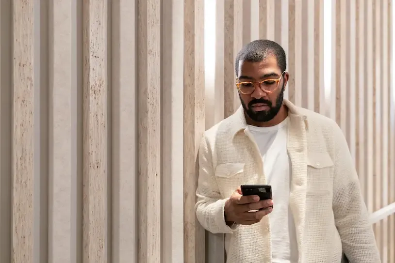 A man with a beard and glasses stands against a wall with vertical wooden slats. He is wearing a white t-shirt, a cream-colored textured button-down shirt, and black pants. He is looking down at a smartphone in his hands and holding a laptop under his arm.