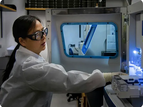 A scientist in a white lab coat and safety goggles focused on operating a piece of high-tech laboratory equipment with a blue-rimmed viewing window.
