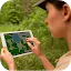Side view of a woman in a khaki uniform and cap using a digital tablet to view a topographic map while standing in a lush, green wooded area.