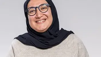 A smiling woman with glasses wearing a black hijab and a beige knit sweater, posed against a neutral grey background for a professional headshot.