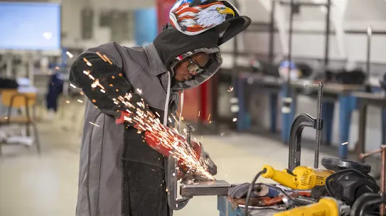 A young person wearing a welding helmet with an American flag and eagle design, protective glasses, and a grey jacket with leather sleeves is using an angle grinder on a metal piece at a workbench, creating a shower of bright orange sparks.