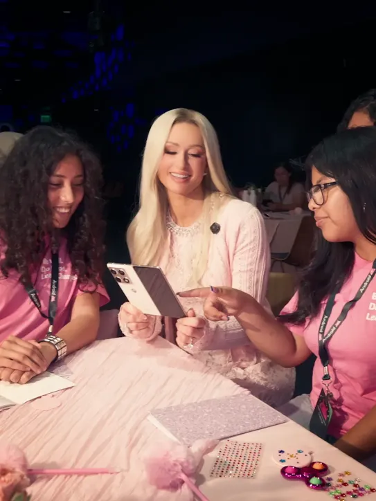 Paris Hilton sitting at a pink table with two girls in pink shirts, holding a folding smartphone while the girl on the right points at the screen.