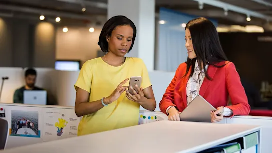 Two professional women collaborating in an office; one is using a smartphone while the other holds a tablet and looks on.