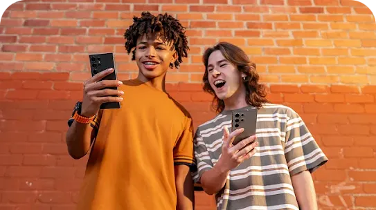 Zwei junge Erwachsene halten Android Smartphones in der Hand und machen ein Selfie vor einer roten Backsteinwand – moderne Handy-Nutzung im Alltag.