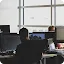 A rear view of a developer working at a computer desk in a bright, modern office. Multiple monitors show lines of code and technical diagrams, with a large window in the background.