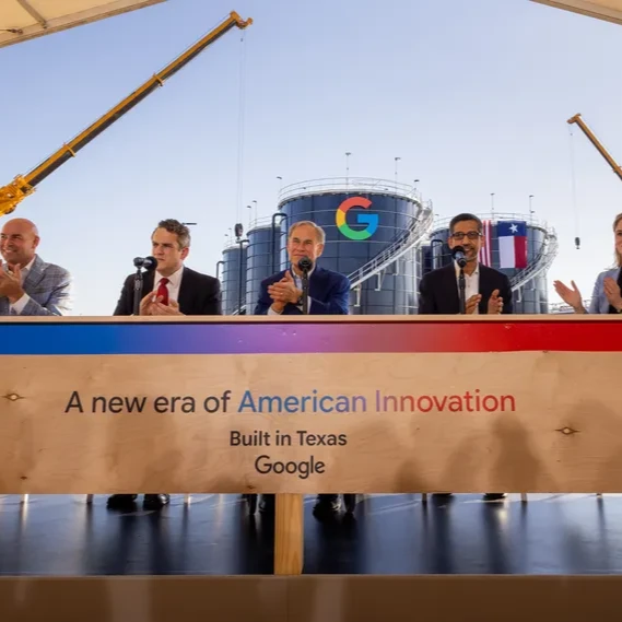 Branded Google event podium against industrial storage tanks bearing Texas flags, flanked by cranes.