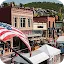 A high-angle view of a bustling historic American main street featuring brick buildings, outdoor cafe seating, and an American flag in the foreground, with mountain houses in the background.