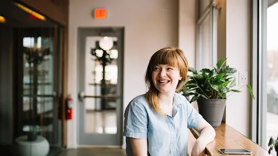 A smiling woman sitting at a wooden counter in a brightly lit cafe or office space.