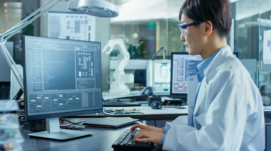 Asian Scientist Sitting at His Desk Doing Sophisticated Coding and Programming on His Desktop Computer. In the Background Computer Science Research Laboratory with Robotic Arm Model.