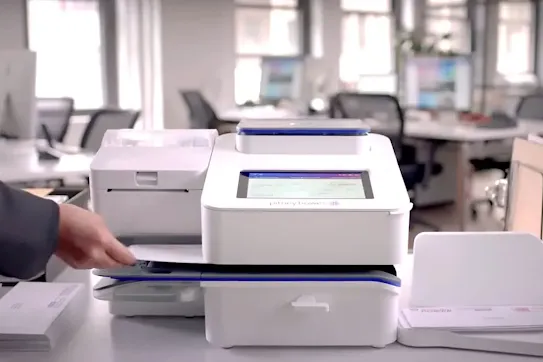 A person an envelope into a white Pitney Bowes digital postage meter and mailing machine in an office.