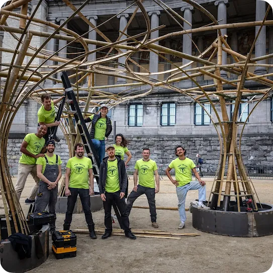 A group of eight construction team members wearing matching neon green t-shirts featuring "The Trilix" logo pose in front of and within a large, intricate bamboo structure installation. Some members stand on a ladder while others pose on the ground near toolboxes, with a classical stone building featuring large pillars in the background.