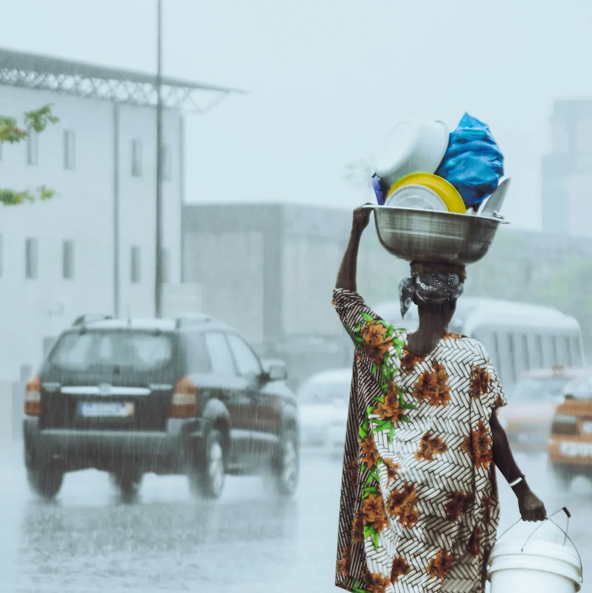 A woman in a patterned dress balances a bowl of dishes on her head while walking in a downpour on a city street.