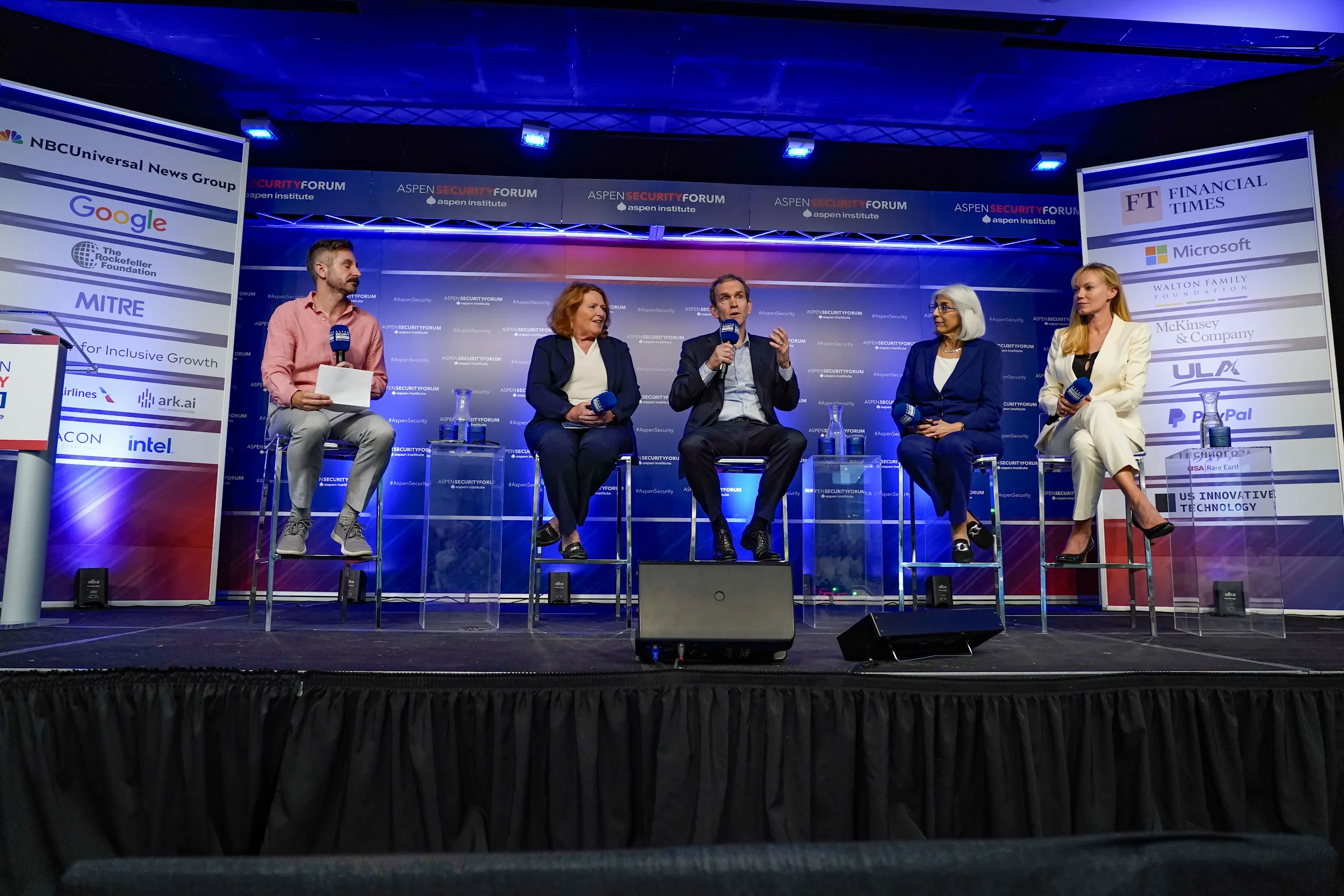 A wide-shot of a five-person panel discussion on a stage at the Aspen Security Forum. The stage is flanked by tall banners featuring logos from Google, NBCUniversal, Financial Times, Microsoft, and other sponsors. The diverse group of experts is seated on high stools against a blue lit backdrop, with a moderator on the far left leading the conversation.