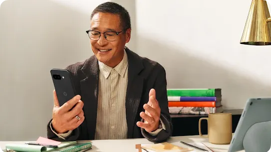 A smiling man wearing glasses holds a smartphone while working at a desk with a tablet and notebooks.
