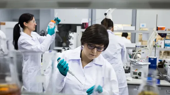 A female scientist using a pipette in a Ryvu Therapeutics laboratory, with other researchers working in the background.