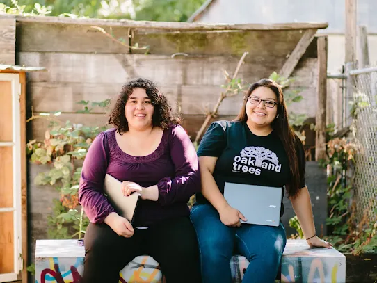 Two smiling women sitting side-by-side on a graffiti-painted bench outdoors while holding closed laptops. The woman on the left wears a purple shirt and black pants, and the woman on the right wears glasses, blue jeans, and a black t-shirt that says "oakland trees." They are positioned in front of a rustic wooden fence and garden greenery.
