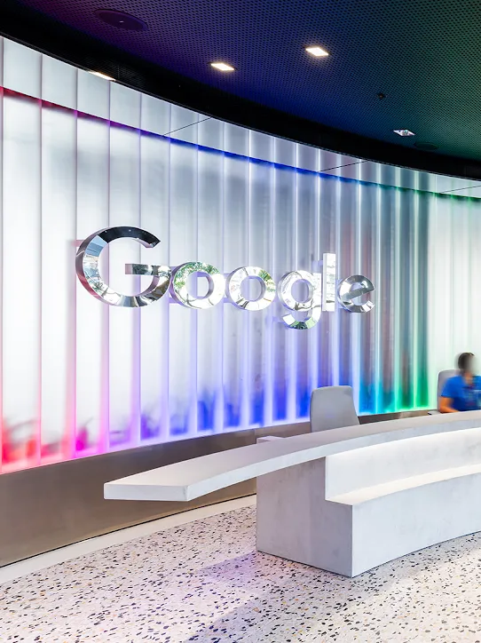 Red, blue and green lighting illuminate a curved wall with a silver Google logo. In front, a person sits behind a white desk, with three people passing by.
