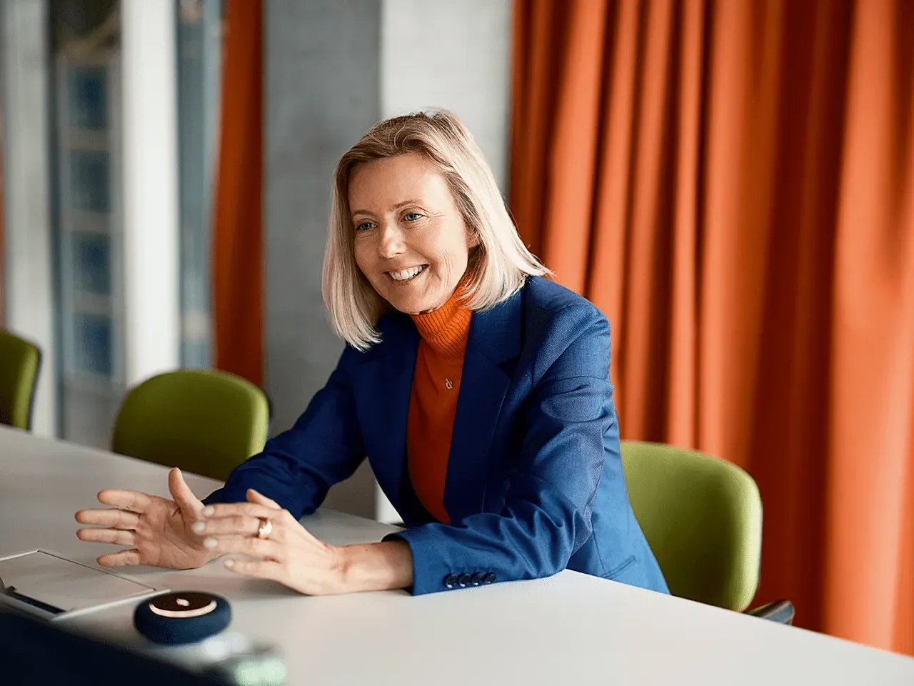 Dr. Marianne Janik, CEO of Microsoft Germany, speaking during an interview at the Google office in Zurich. She is wearing a blue blazer and orange turtleneck, gesturing with her hands while seated at a conference table against a background of orange curtains.