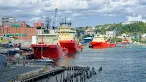 Several large red and yellow industrial offshore supply vessels are docked at a city port, with a hillside covered in colorful buildings in the background.