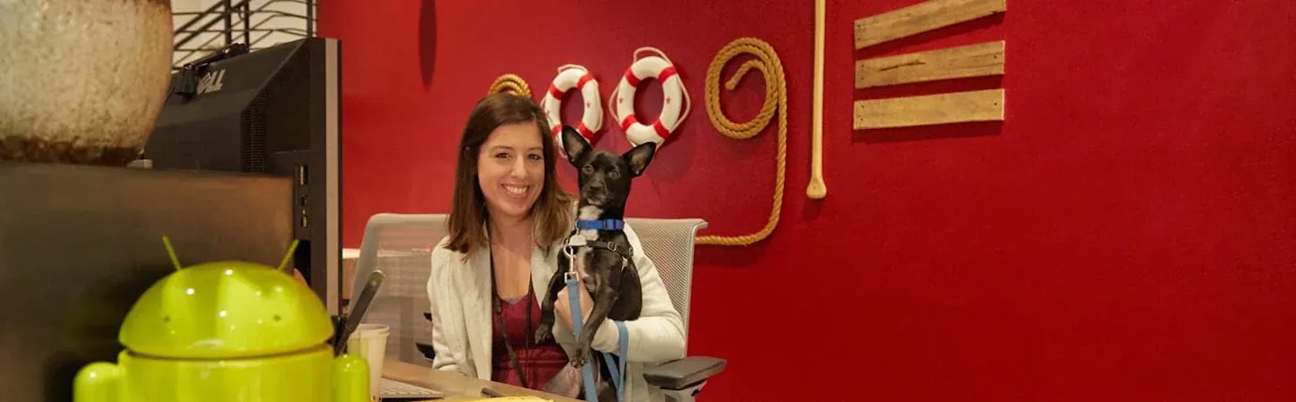 Woman sitting at a desk holding a small black dog, with a green Android figure in the foreground and nautical decor on a red wall in the background