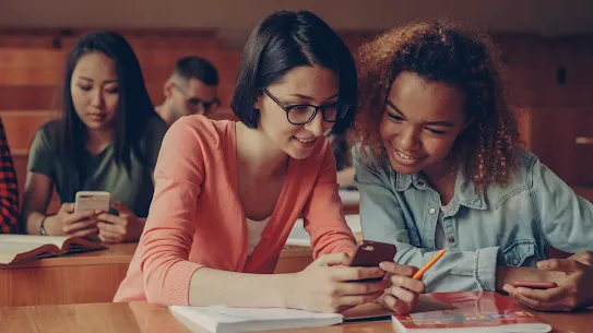 Two young female college students sitting at a wooden desk in a lecture hall, smiling while looking at a smartphone together during class.