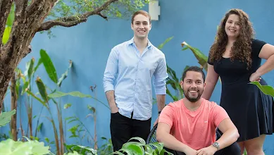 Cuidas founders João, Matheus, and Deborah pose together in front of a blue wall.