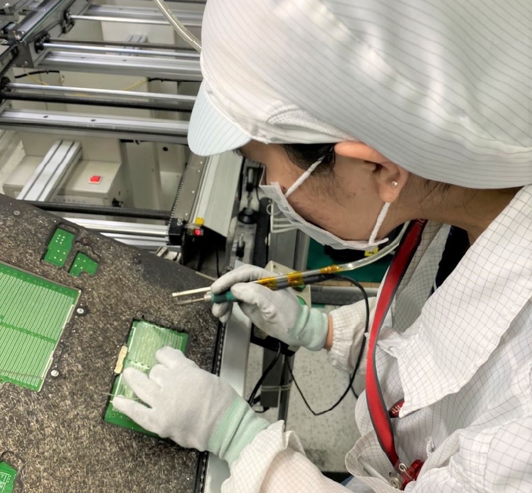 A supplier wearing white gloves, a checkered shirt, striped head covering and face mask while handling a soldering tool