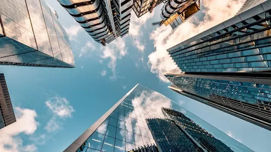 A dramatic low-angle perspective looking straight up at several modern skyscrapers reaching toward a blue sky with soft white clouds. The buildings feature diverse architectural styles, including glass-walled towers reflecting the sky and an industrial-style structure with external metallic pipes and scaffolding. This visual represents the "AI Sprinters" report and the building of modern government infrastructure.