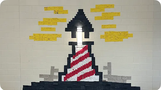 A wall display featuring a lighthouse created from arranged sticky notes, incorporating a light fixture as the beacon, flanked by yellow sticky notes shaped like clouds containing handwritten motivational messages.