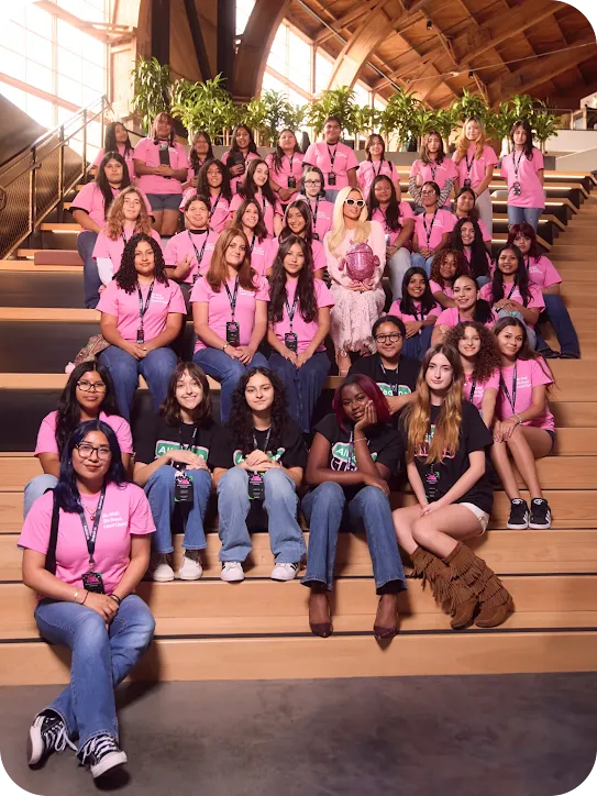 Group photo on wooden bleachers featuring Paris Hilton holding a pink Android mascot, surrounded by dozens of smiling girls in pink and black shirts.