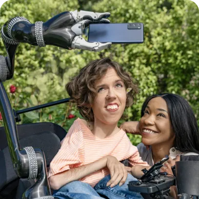 A white person with wavy brown hair sits in their motorized wheelchair with a robot arm attachment holding an Android phone. A Black woman crouches next to them and smiles up at the Android phone.